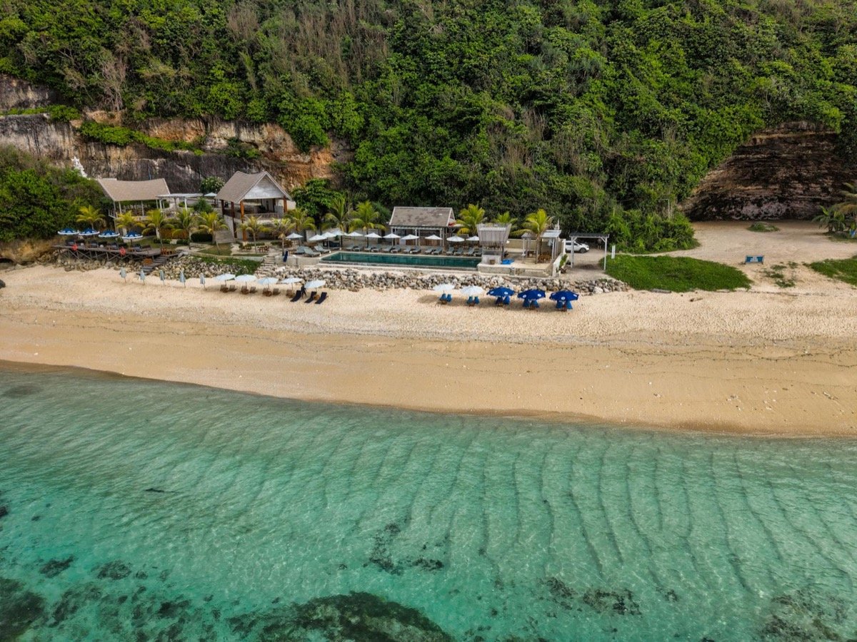 Vista aérea de un resort de lujo junto al mar con aguas turquesas, playa de arena y exuberante colina tropical de fondo