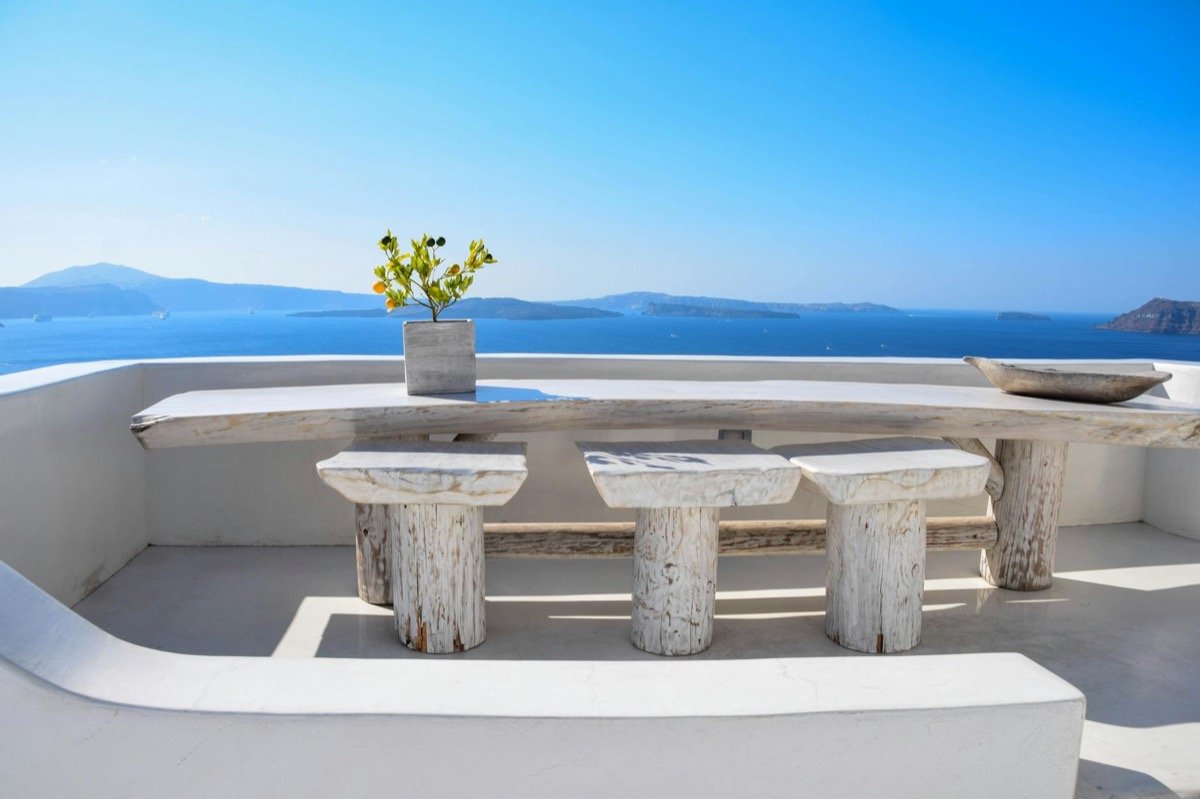 Rustic stone dining table with wooden stools on a whitewashed terrace overlooking the Santorini caldera and Aegean Sea under a clear blue sky