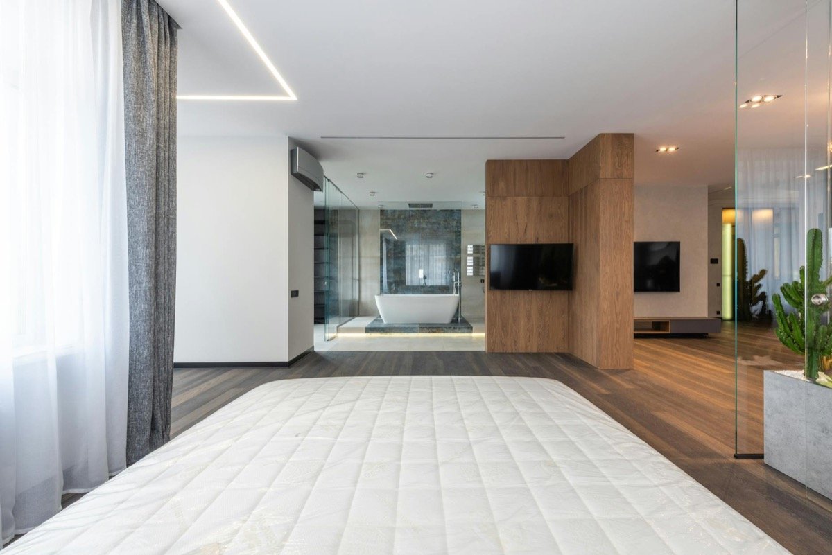 Spacious modern luxury hotel suite with minimalist white decor, king bed, freestanding bathtub behind glass partition, and floor-to-ceiling windows