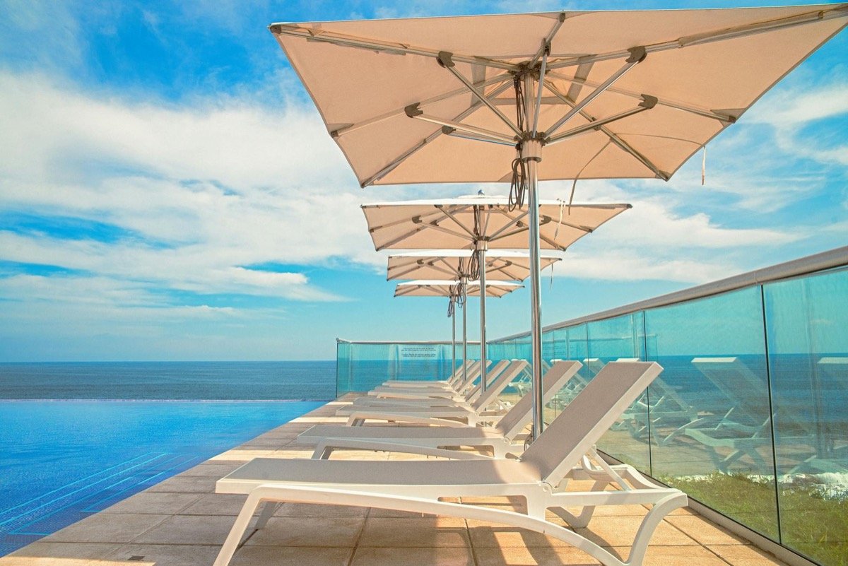 Luxury poolside sun loungers with parasols lined up along an infinity pool edge overlooking the deep blue sea on a sunny day